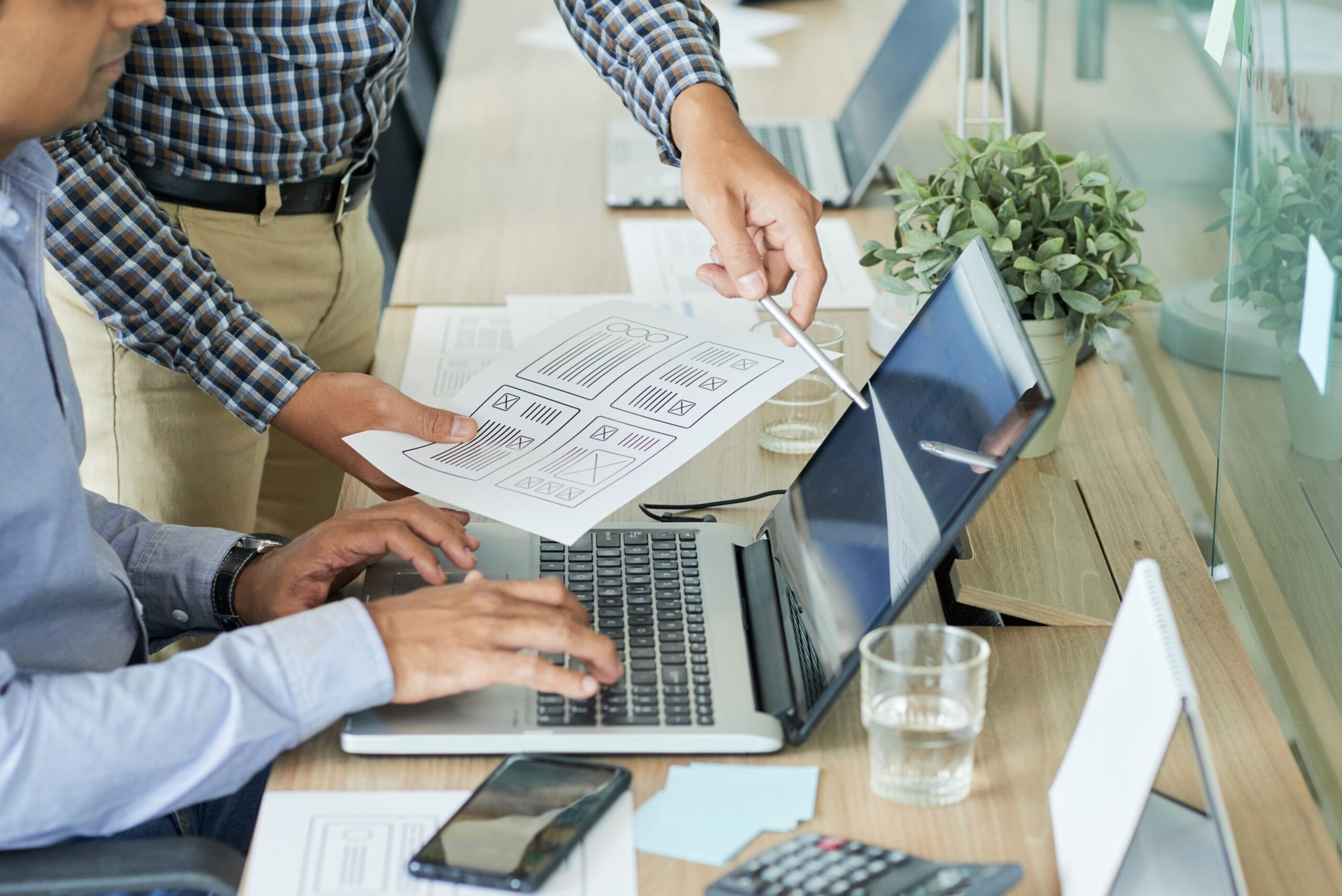 Virtual admin assistance services — Two men in front of an office desk with a laptop on it, analyzing the printed design for a website to be developed.